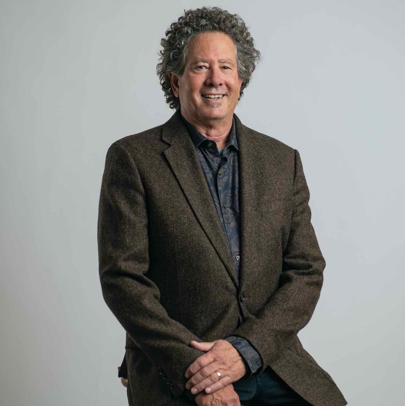Studio portrait of Barry Brown with curly gray hair, wearing a brown blazer over a dark patterned shirt against a light gray background.